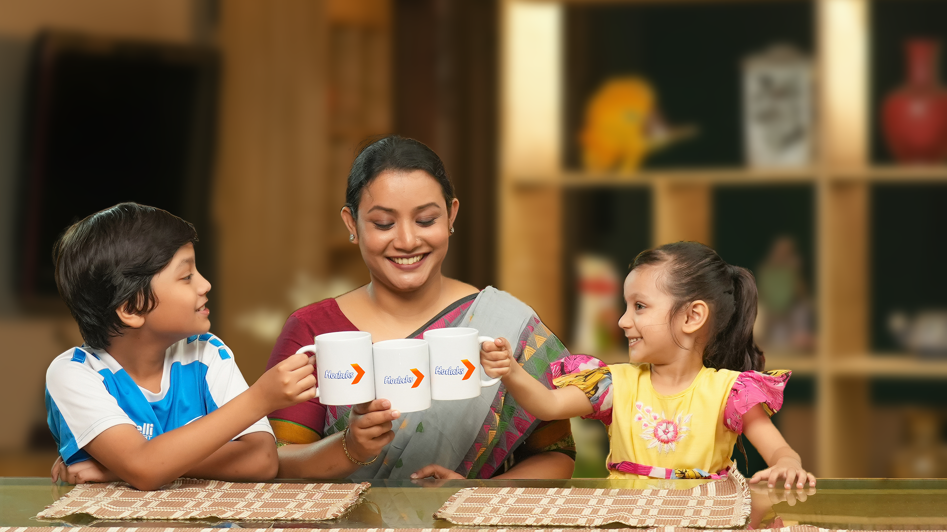 A smiling woman and two children raise mugs in a cozy kitchen.