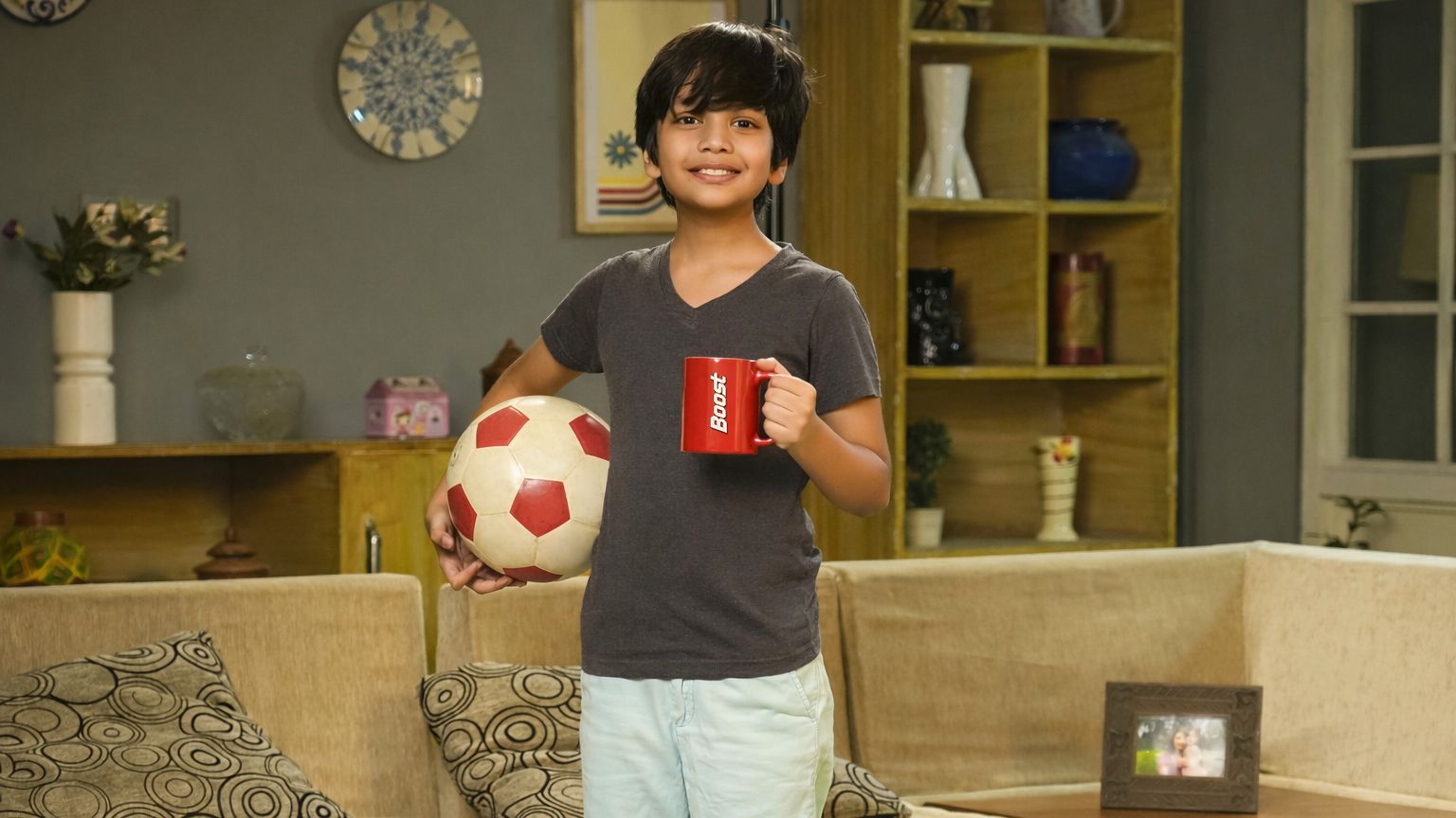 A young boy stands in a cozy living room, smiling, holding a red coffee mug and a soccer ball.
