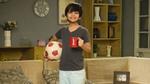 A young boy stands in a cozy living room, smiling, holding a red coffee mug and a soccer ball.