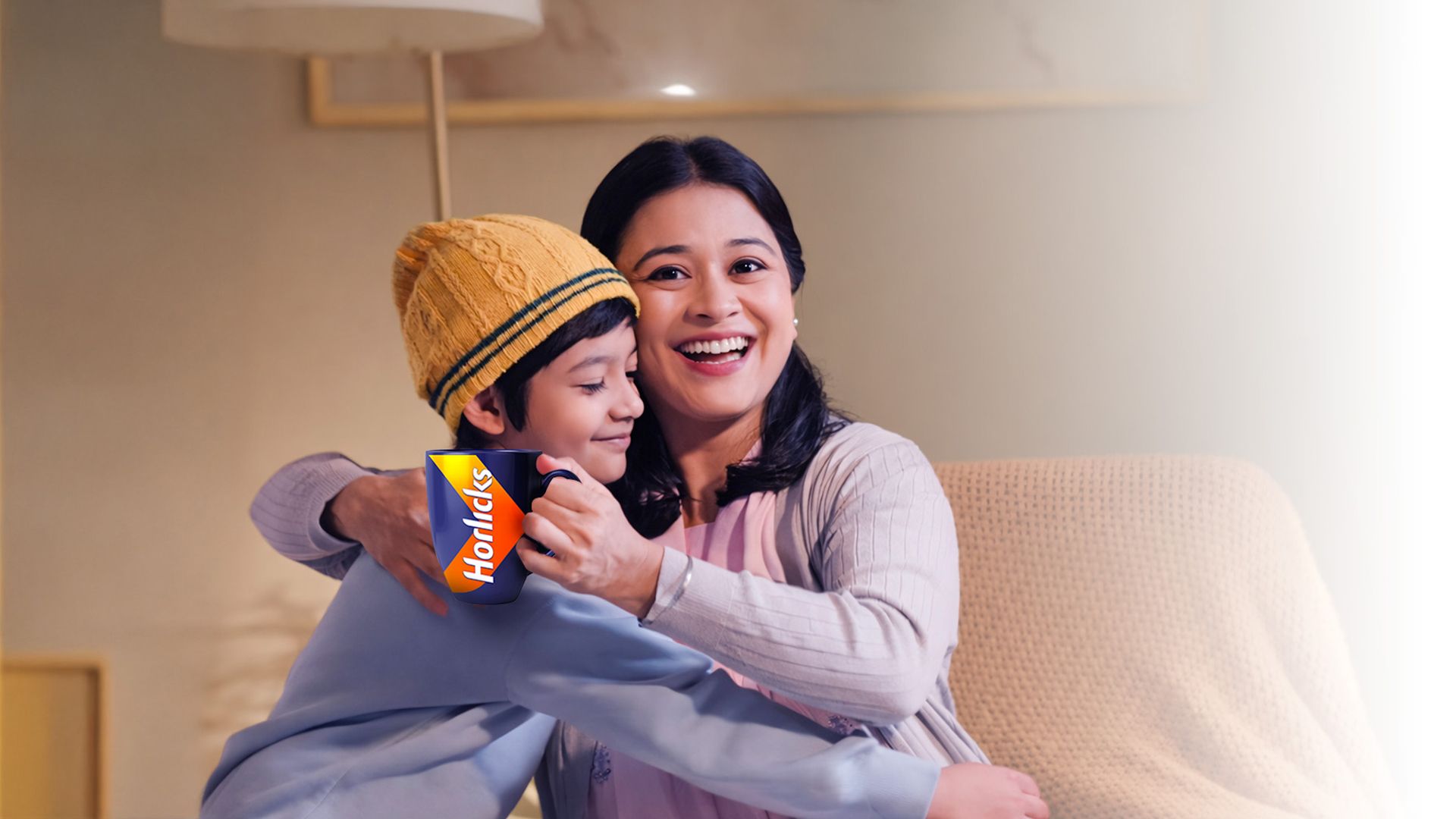 A smiling woman hugs a child in a yellow hat holding a Horlicks packet, sitting on a cozy chair.