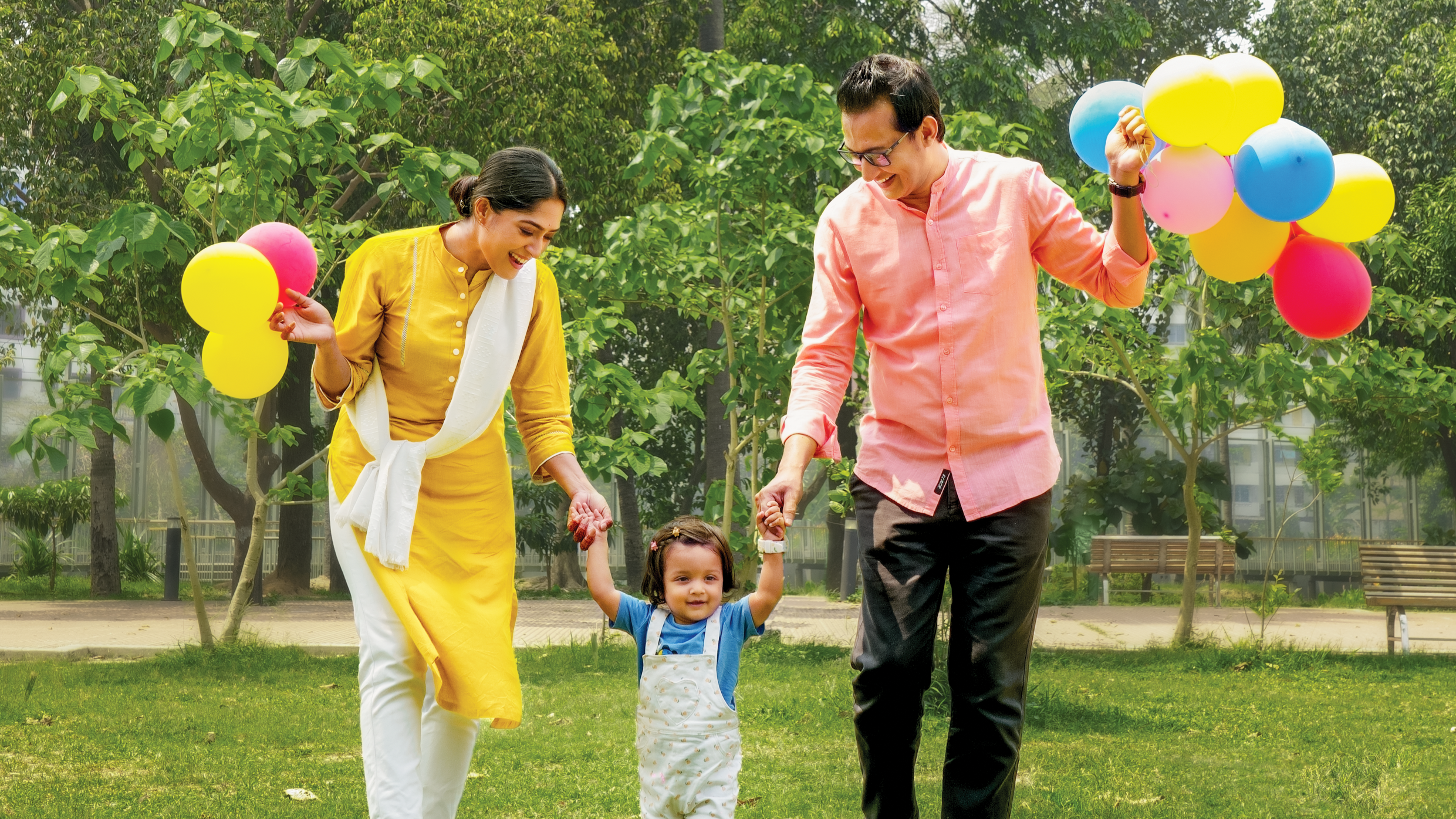 A joyful family of three walks in a park, parents holding colorful balloons and assisting their smiling child in the center. Green trees and grass in the background.